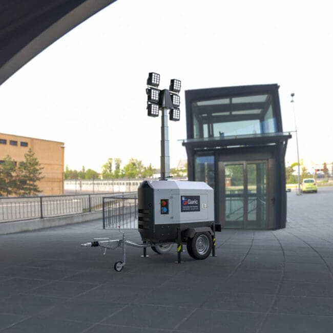 The Eco Tower Light, equipped with four large lights, is positioned on a paved outdoor area next to a modern glass building.