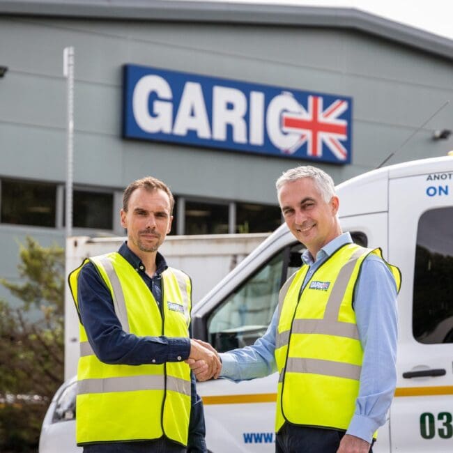 Two men in yellow safety vests shake hands outside a building with a Garic sign and a van.