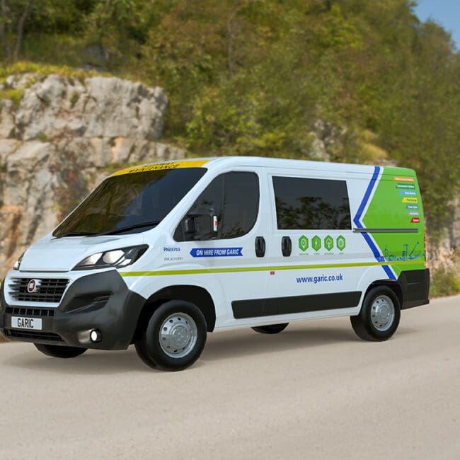 The Welfare Van Eco, a white electric van with colorful graphics, drives along a rural road bordered by rocky hills and green trees.