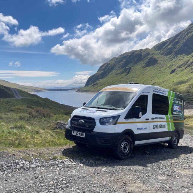 A white van parked on a gravel road by a lake, surrounded by green hills under a partly cloudy sky.