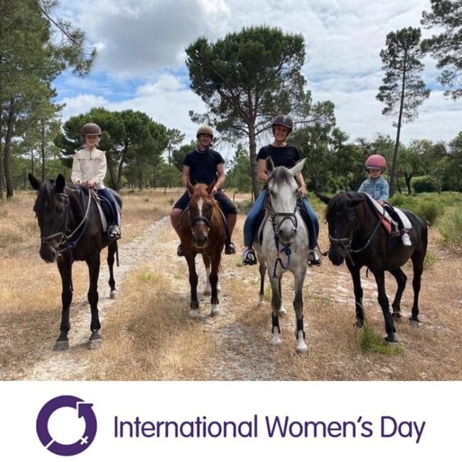 Four people riding horses on a path, with trees in the background and an International Womens Day logo below.