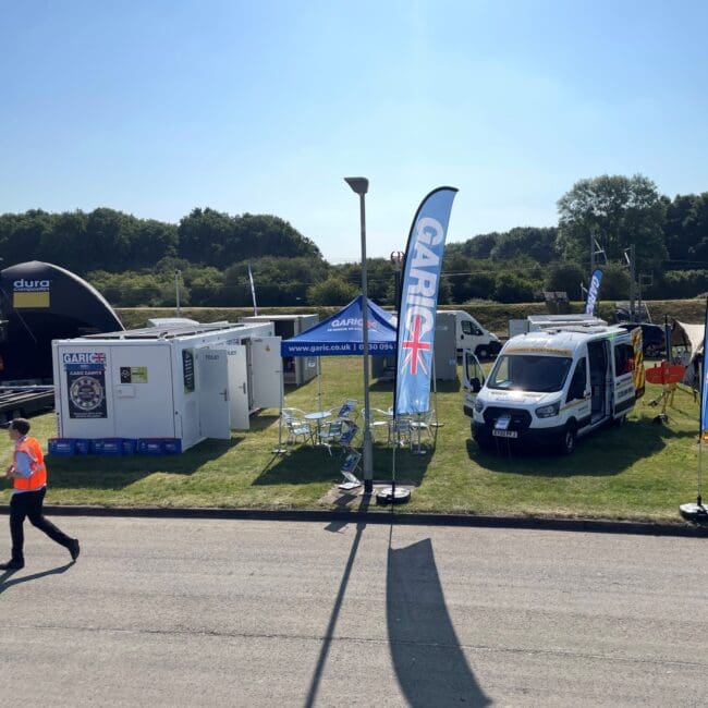 Outdoor event setup with GARIC banners, tents, vehicles, and people on a sunny day.