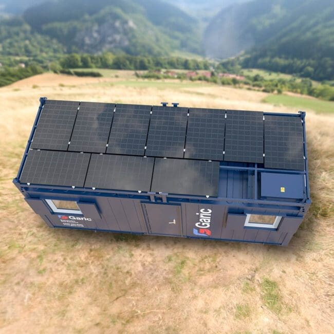 The Solar Power Frame sits in a grassy field with hills and mountains behind it, featuring solar panels installed on the roof of its blue container.