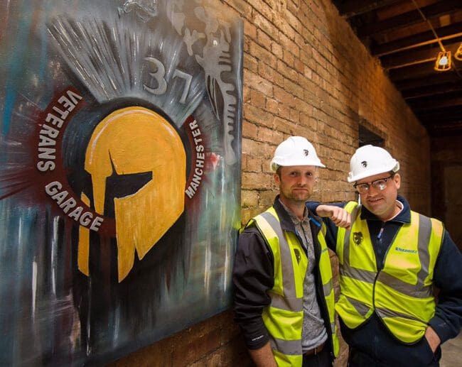 Two men in hard hats and vests stand beside a Veterans Garage Manchester mural on a brick wall.