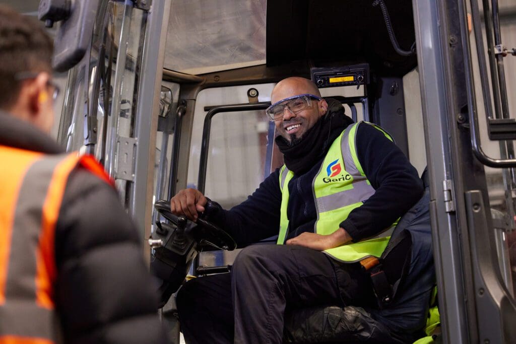 Man in safety gear smiles while sitting in a construction vehicle, talking to a coworker in an orange vest.