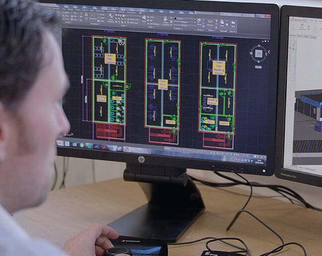 A person works on architectural floor plans displayed on dual computer monitors using design software.