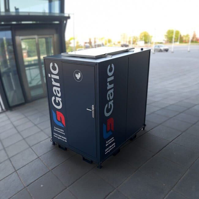 A Solar Loo 8 portable toilet unit, in dark blue, stands on a paved outdoor area near a modern glass building.