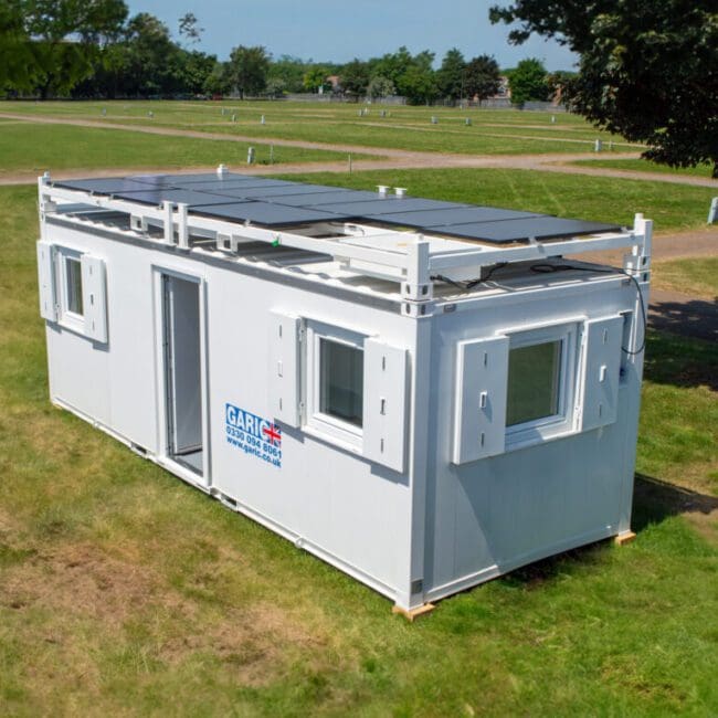 White portable container unit with rooftop solar panels, placed on grass in an open, sunny park area.