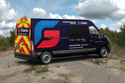 A blue Garic hybrid welfare van is parked outdoors on a dirt road with trees in the background.