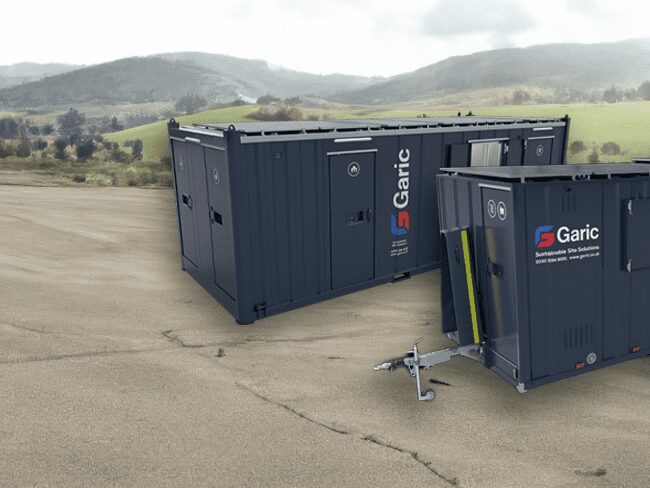 Two dark blue Garic portable cabins on a paved area with green hills and trees in the background.
