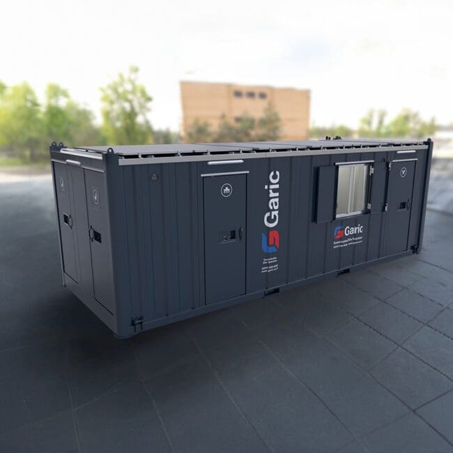 A dark grey Eco Cabin+ 24 portable site cabin by Garic sits on a paved outdoor area with blurred trees and a building in the background.