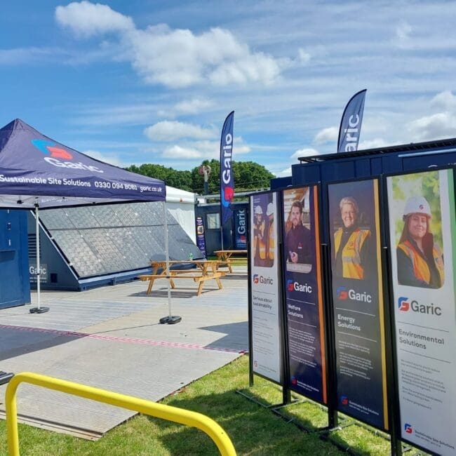 Outdoor display with Garic-branded tents, banners, solar panels, and informational boards on a sunny day.