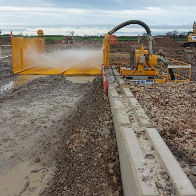 Yellow vehicle wheel wash system spraying water on a muddy construction site with machinery in the background.