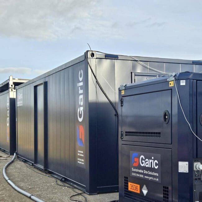 Dark blue Garic portable site cabins and equipment are set up outdoors on a gravel surface.