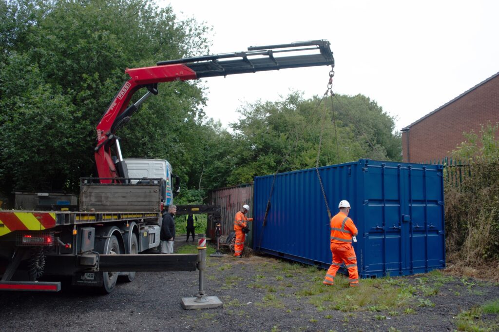 A crane truck lifts a blue shipping container as two workers in orange safety gear stand nearby.
