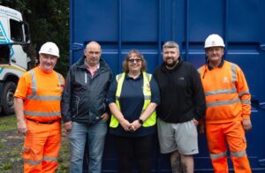 Five people, including two in orange workwear, pose and smile in front of a blue container outdoors.