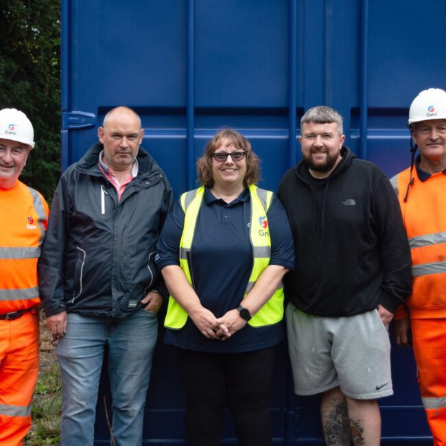 Five people, including two in orange workwear, pose and smile in front of a blue container outdoors.