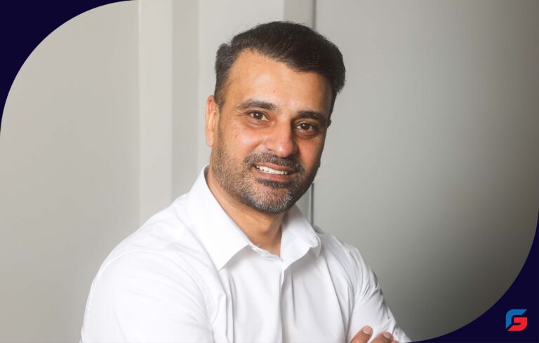Man with short dark hair and beard smiling, wearing a white shirt against a light background.