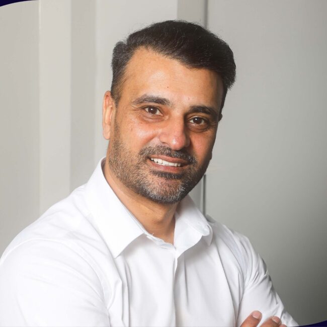 Man with dark hair and beard in white shirt smiling, arms crossed, neutral background.